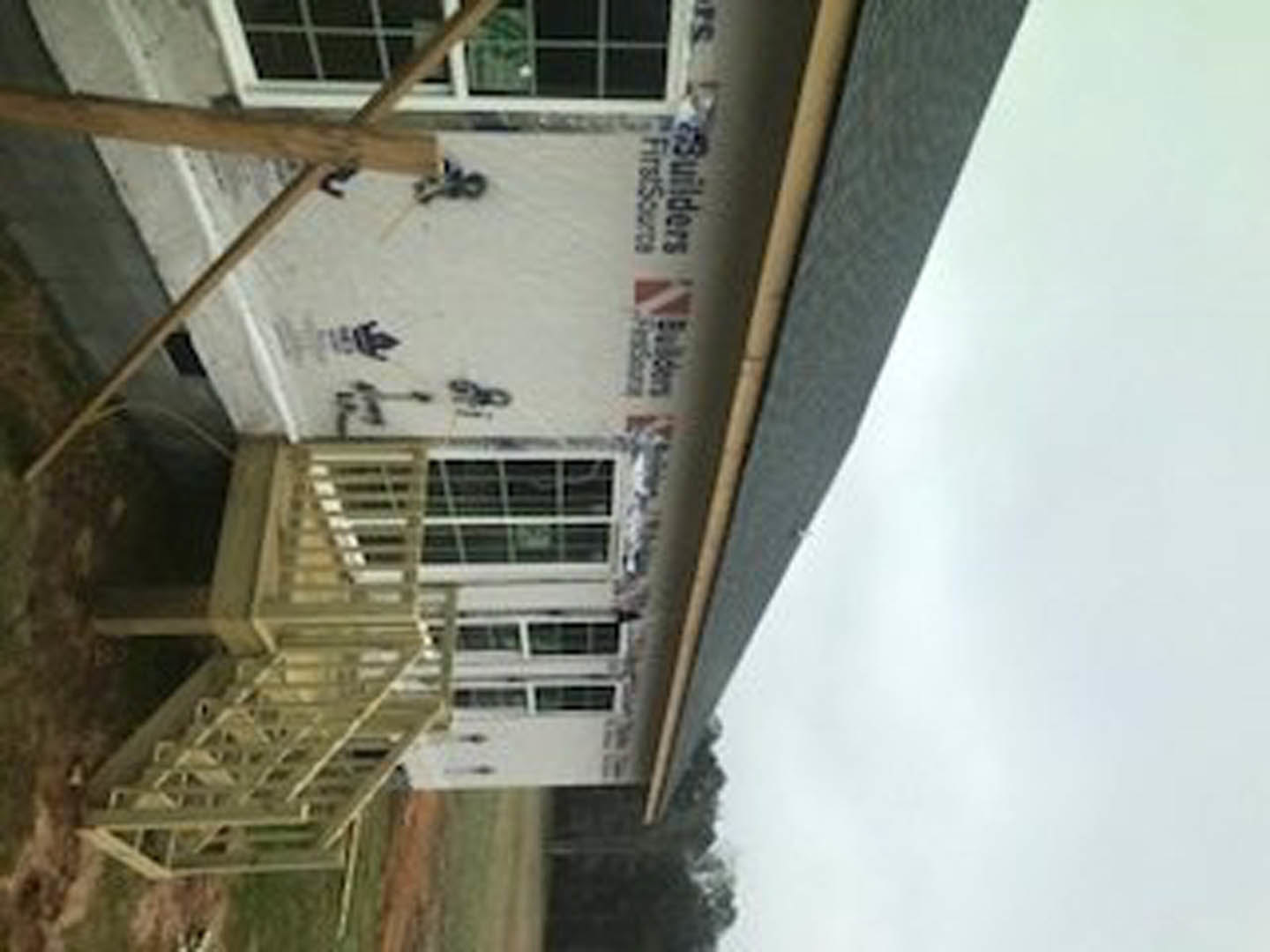 Wood-framed house under construction with exposed deck, metal ladder leaning against exterior, unfinished windows, and partially built wooden stairs.