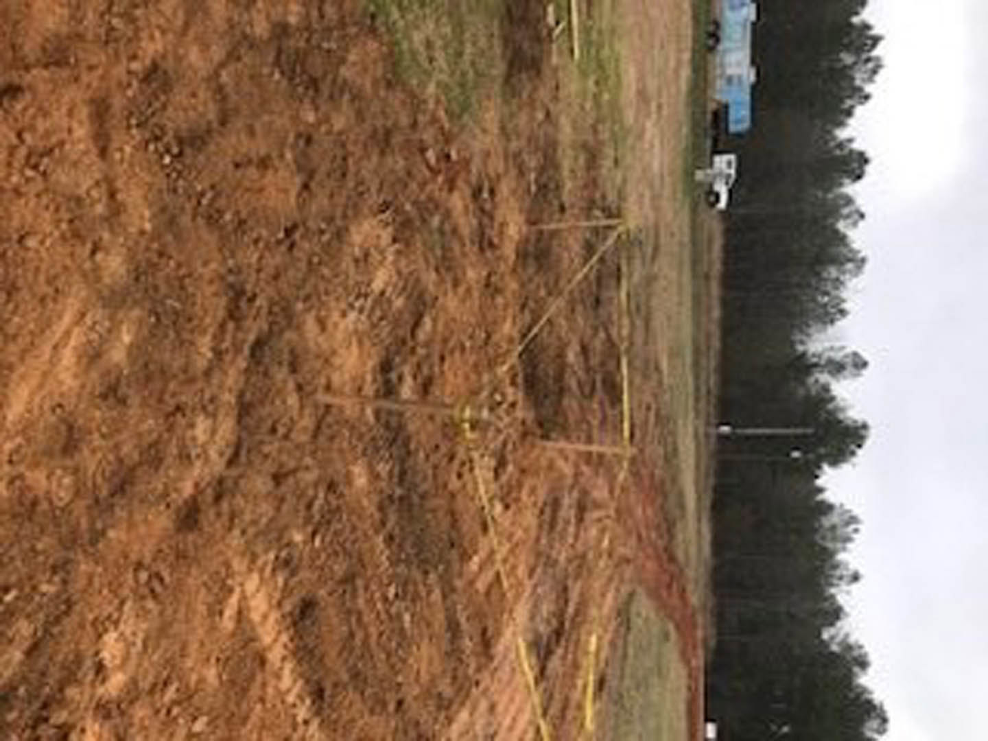Dirt field bordered by trees, scattered yellow poles, patchy grass, and a white truck in the distance