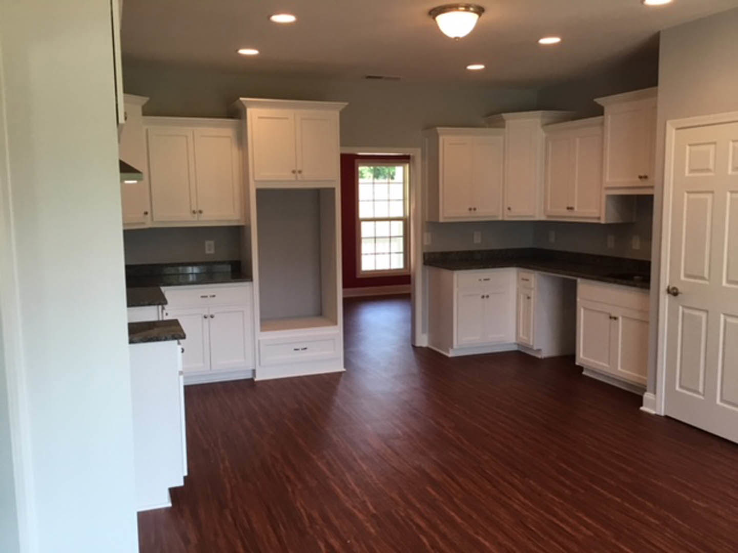 White kitchen cabinets, dark wood flooring, stainless steel appliances, quartz countertops, recessed ceiling lights, white walls, large window, white door with silver hardware