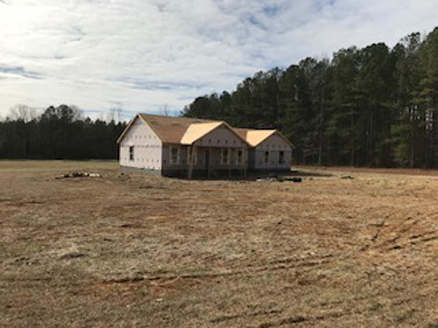 Two-story cottage with gabled roof and large windows, surrounded by grassy field and mature trees under a partly cloudy sky
