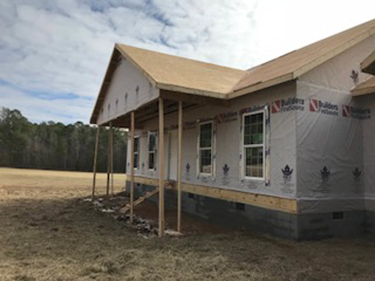 Wood-framed house under construction on grassy lot, white-framed window visible, cloudy sky and trees in background