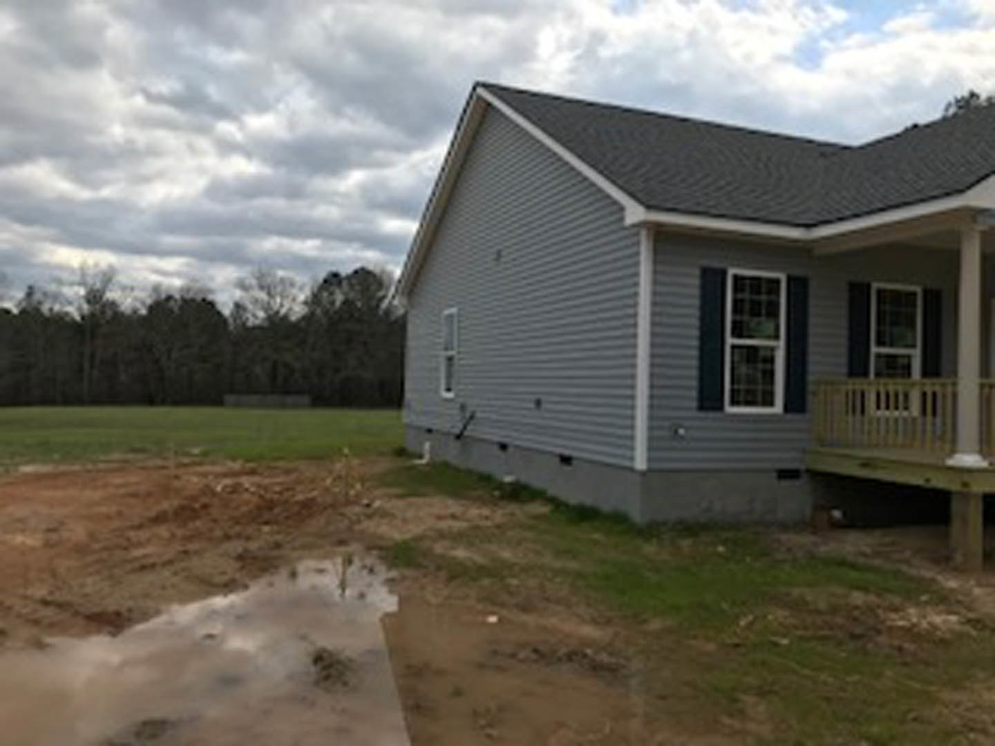 Two-story house with white-framed windows, wood deck and porch railing, puddle of water on dirt in front yard, distant trees, cloudy sky