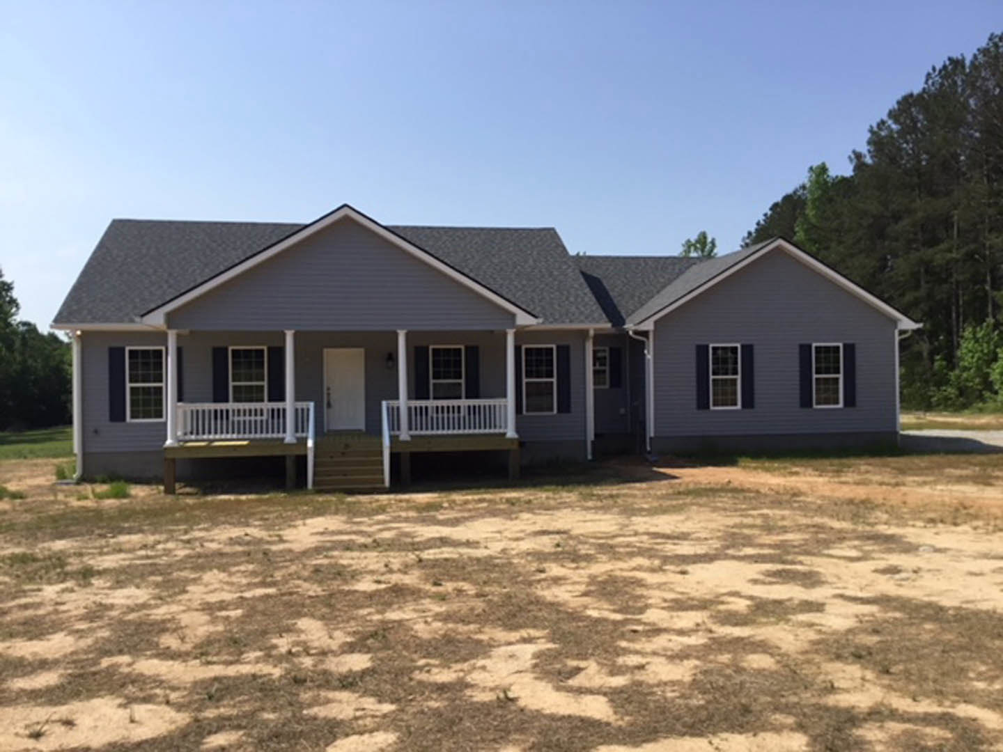 Single-story home with gray siding, covered front porch, grid-style windows, white entry door with black handle, and unfinished dirt yard in foreground