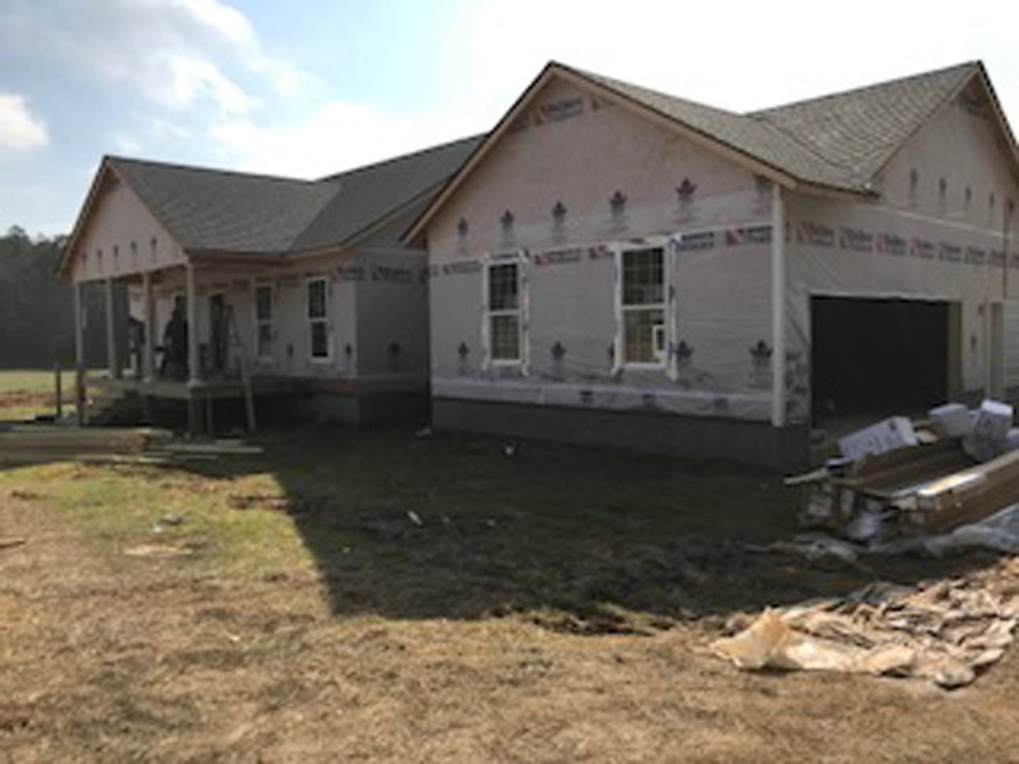 Two-story house under construction with grey roof, white siding, large windows, grassy yard, and pile of construction debris in foreground
