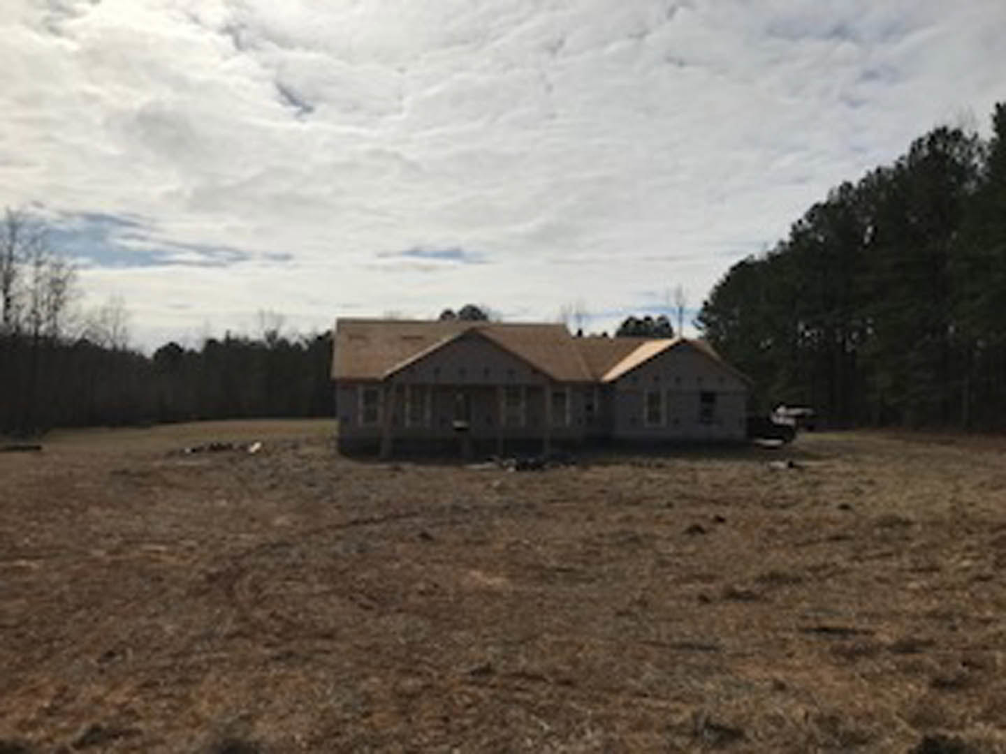 White cottage-style house with a large covered porch, surrounded by grassy field and scattered trees, under a cloudy sky