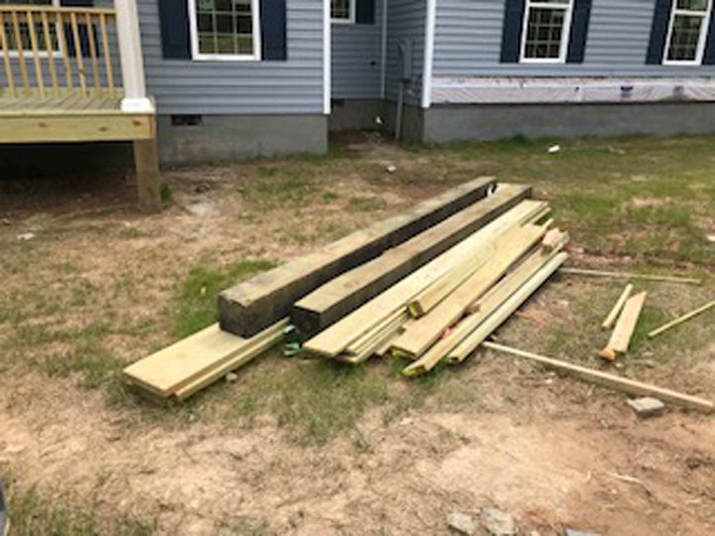 Stack of lumber on grass in front of house with white-framed window and porch, wooden bench nearby