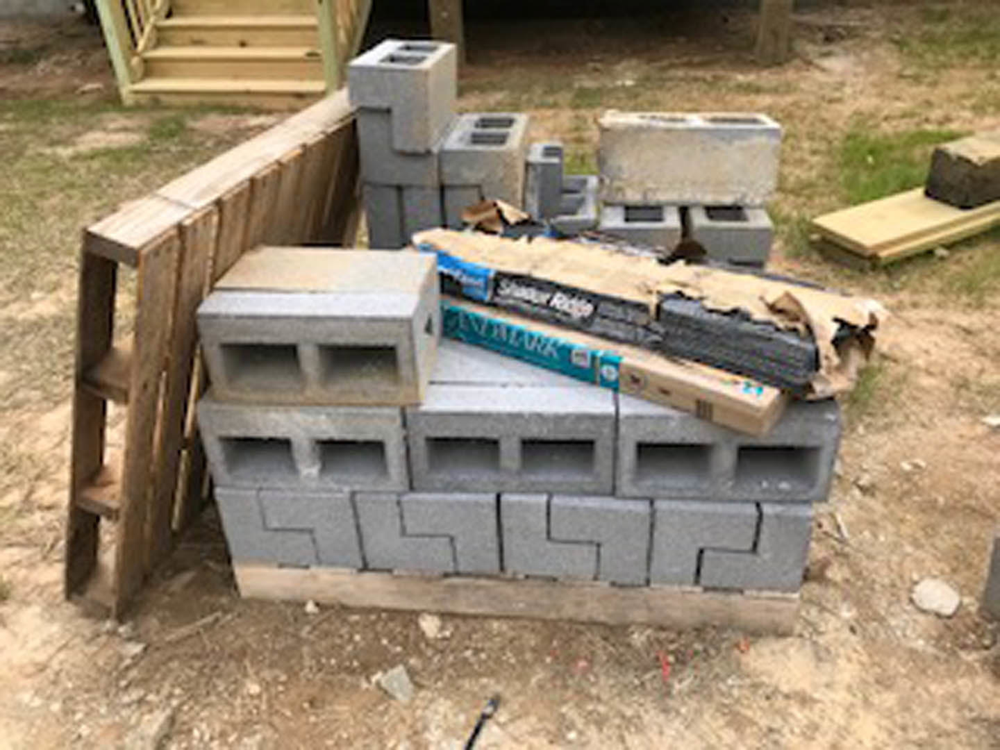 Stack of gray concrete blocks on grassy ground beside unfinished lumber and building exterior