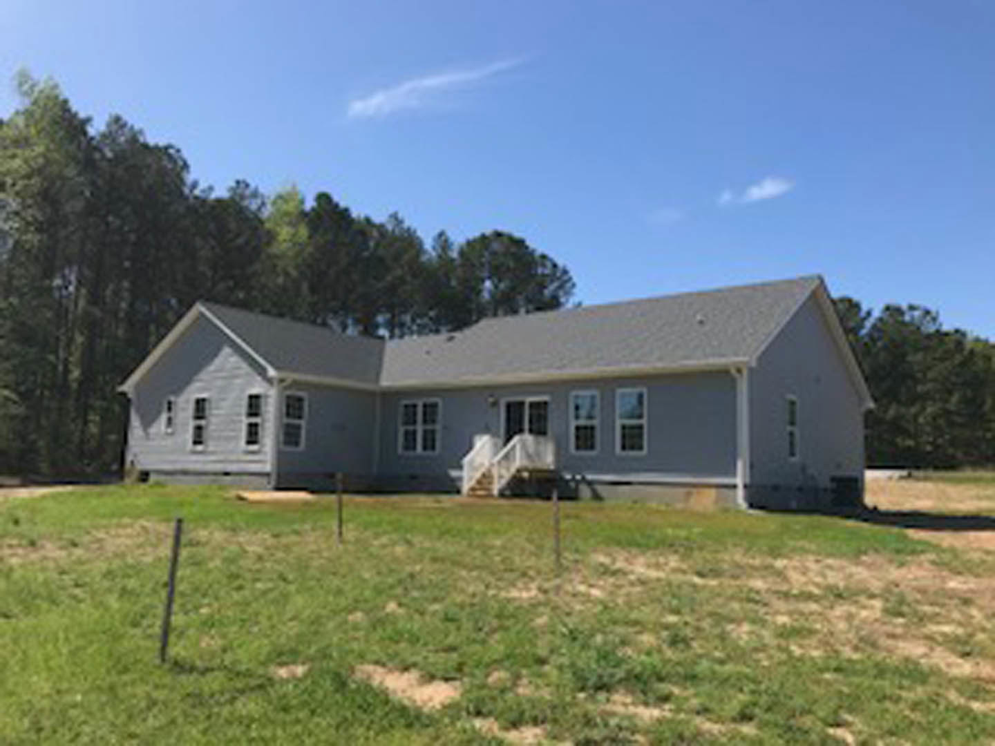 Two-story farmhouse with white siding, surrounded by a wooden fence and expansive green lawn, white-framed windows, and mature trees in the background under a blue sky