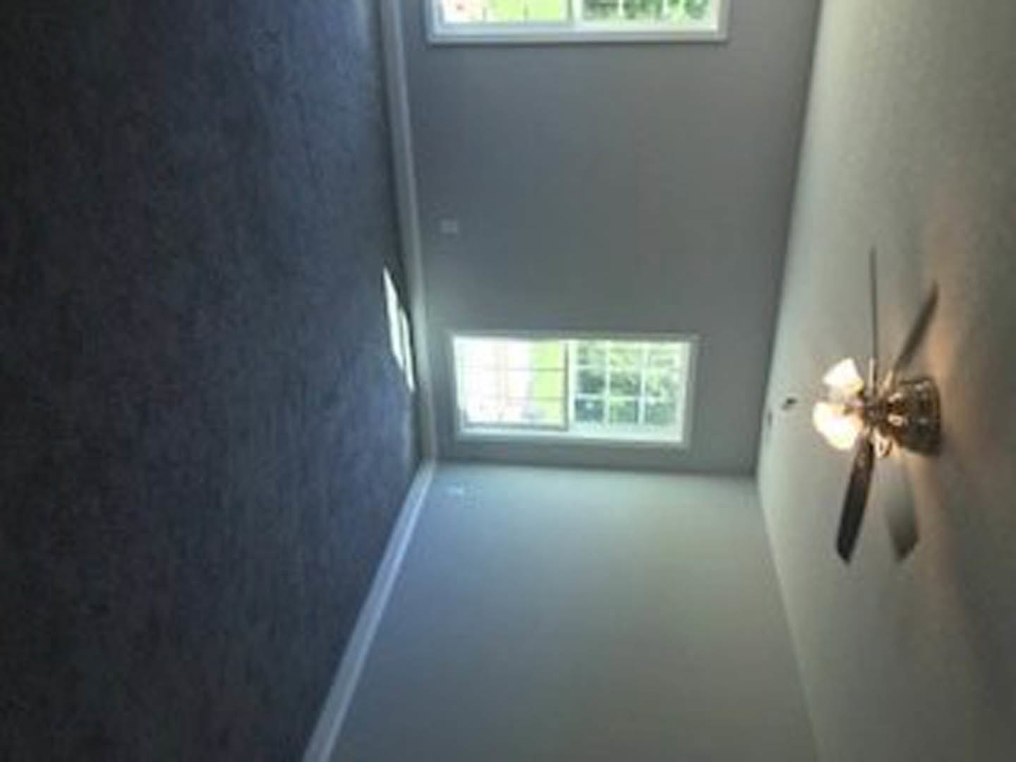 Ceiling fan with brushed metal finish mounted on white ceiling above hardwood floor, large window with outdoor view, blurred foreground.