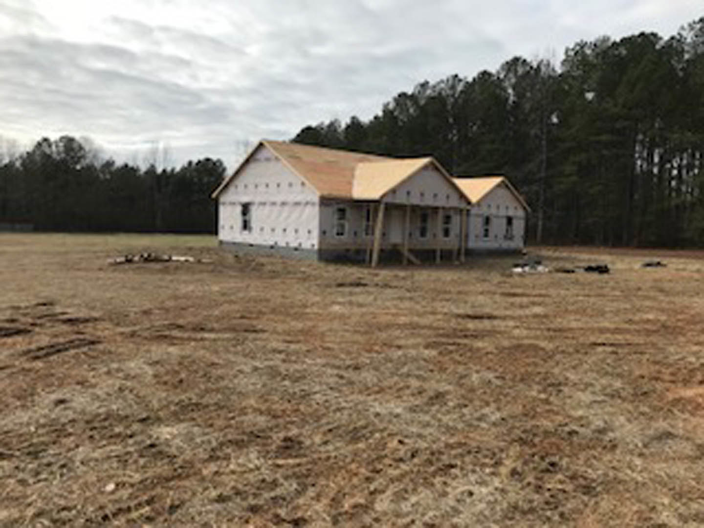 Two-story house with gray shingle roof and white siding set in a grassy field, surrounded by trees under a cloudy sky