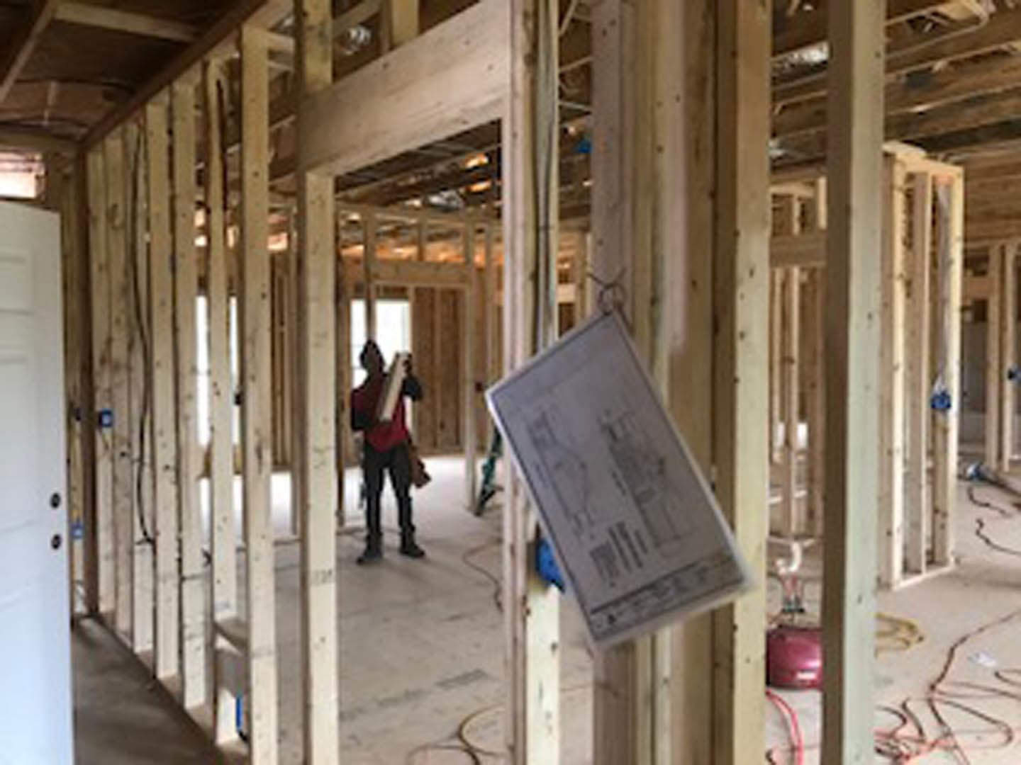 Man in red shirt standing in unfinished room with exposed beams, construction materials, and a blueprint displayed near a doorway with a sign.