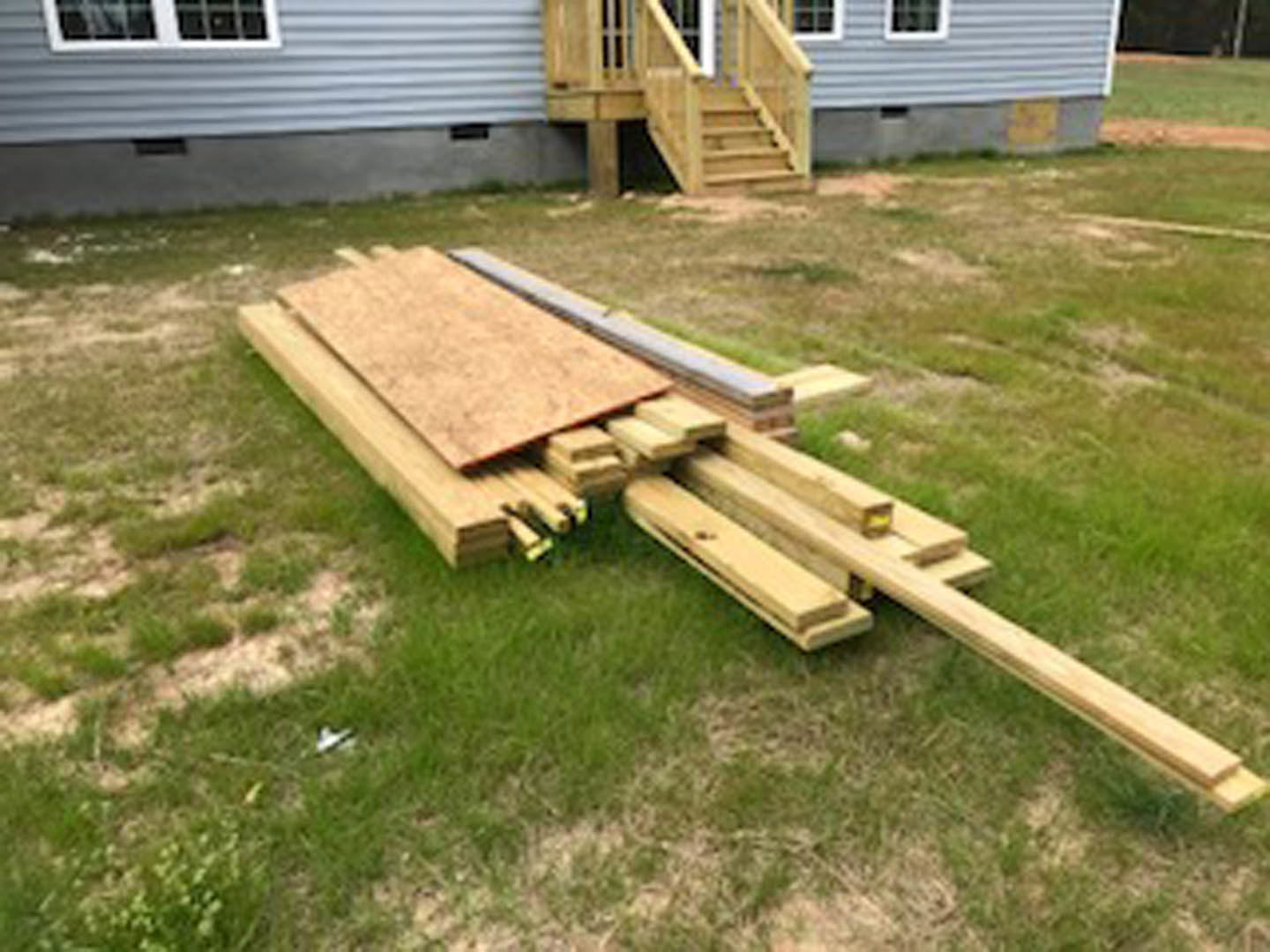 Stack of wooden planks resting on green grass beside a custom home exterior
