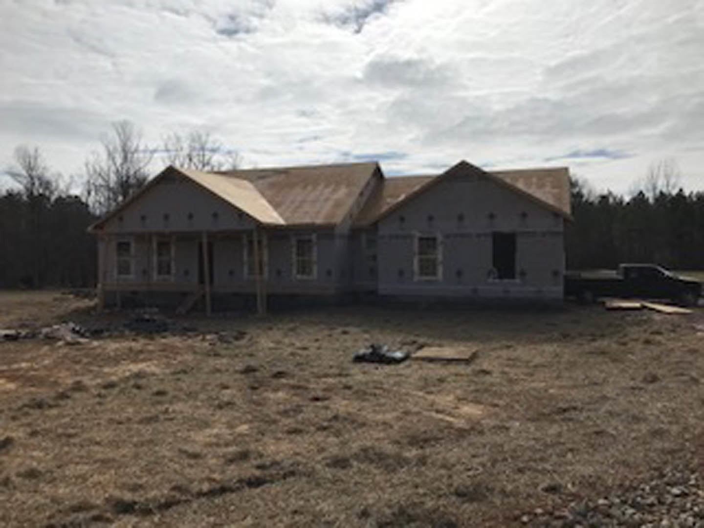 Partially built house with exposed wooden framing and roof, surrounded by dirt field and scattered construction materials, under cloudy sky.