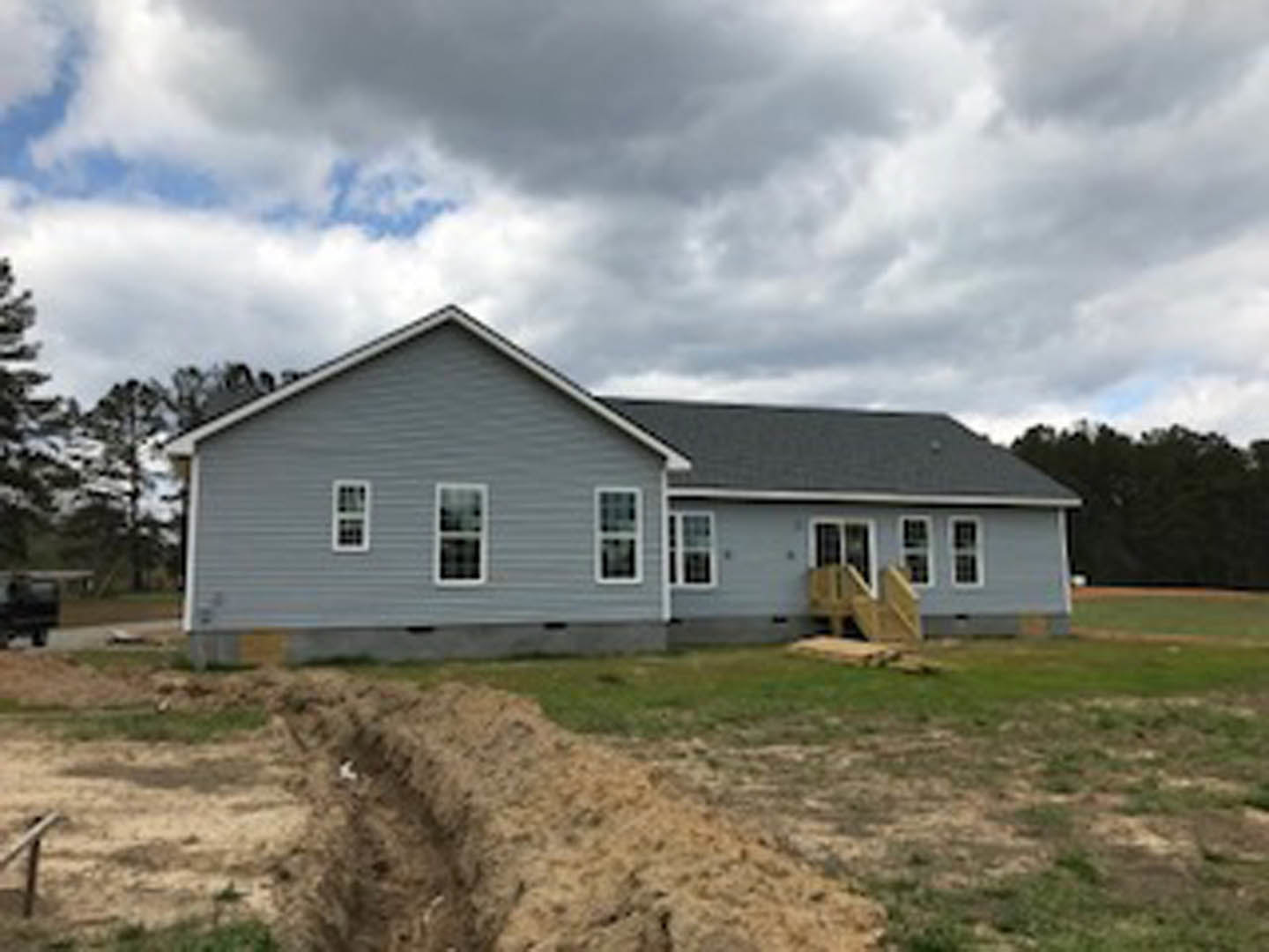 Partially built house with grey roof, white-framed windows, and large dirt excavation in foreground, surrounded by grass and trees under cloudy sky