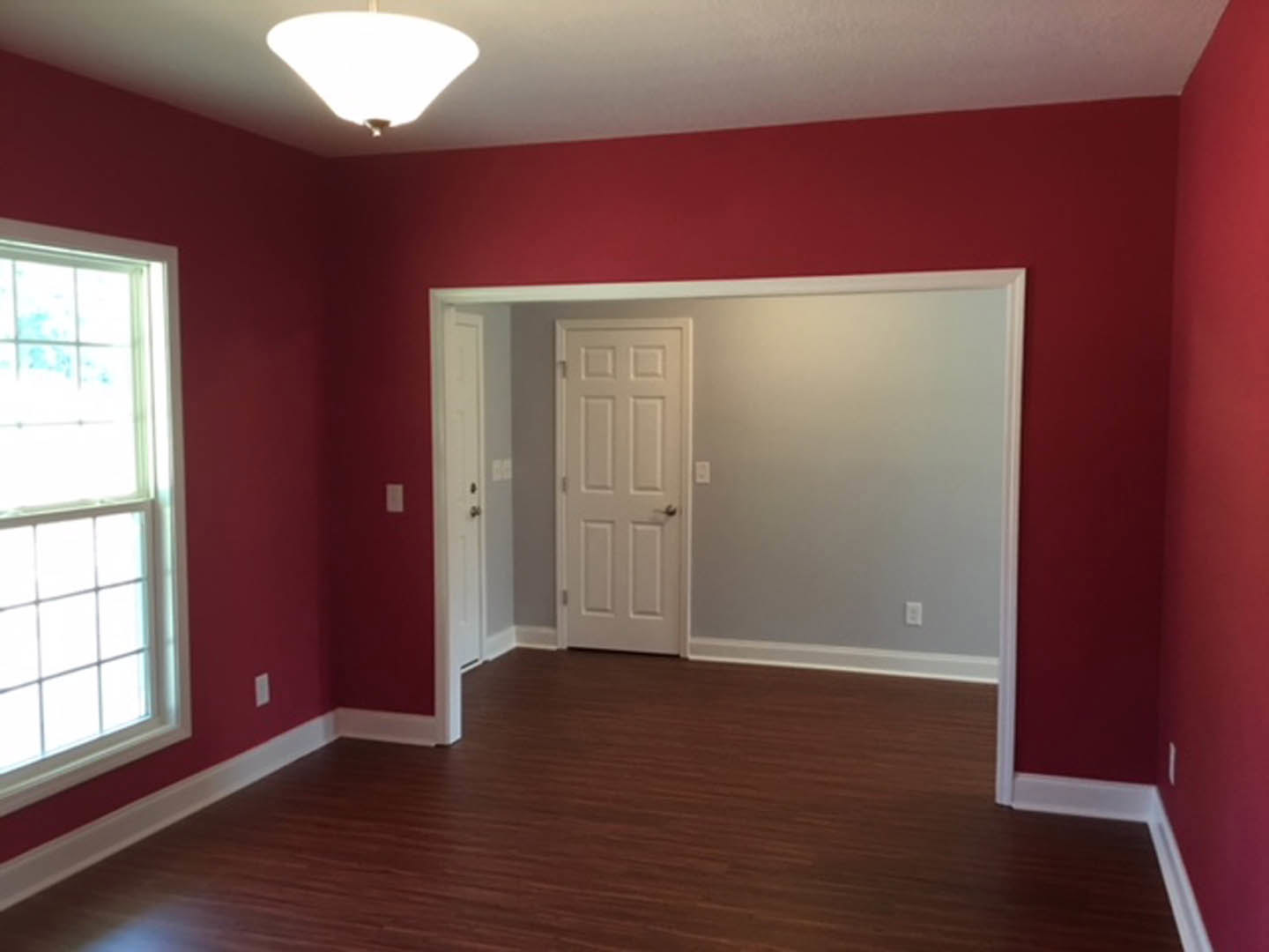 Red walls, wood flooring with white trim, white door featuring a silver handle, window and light fixture visible, plaster ceiling.