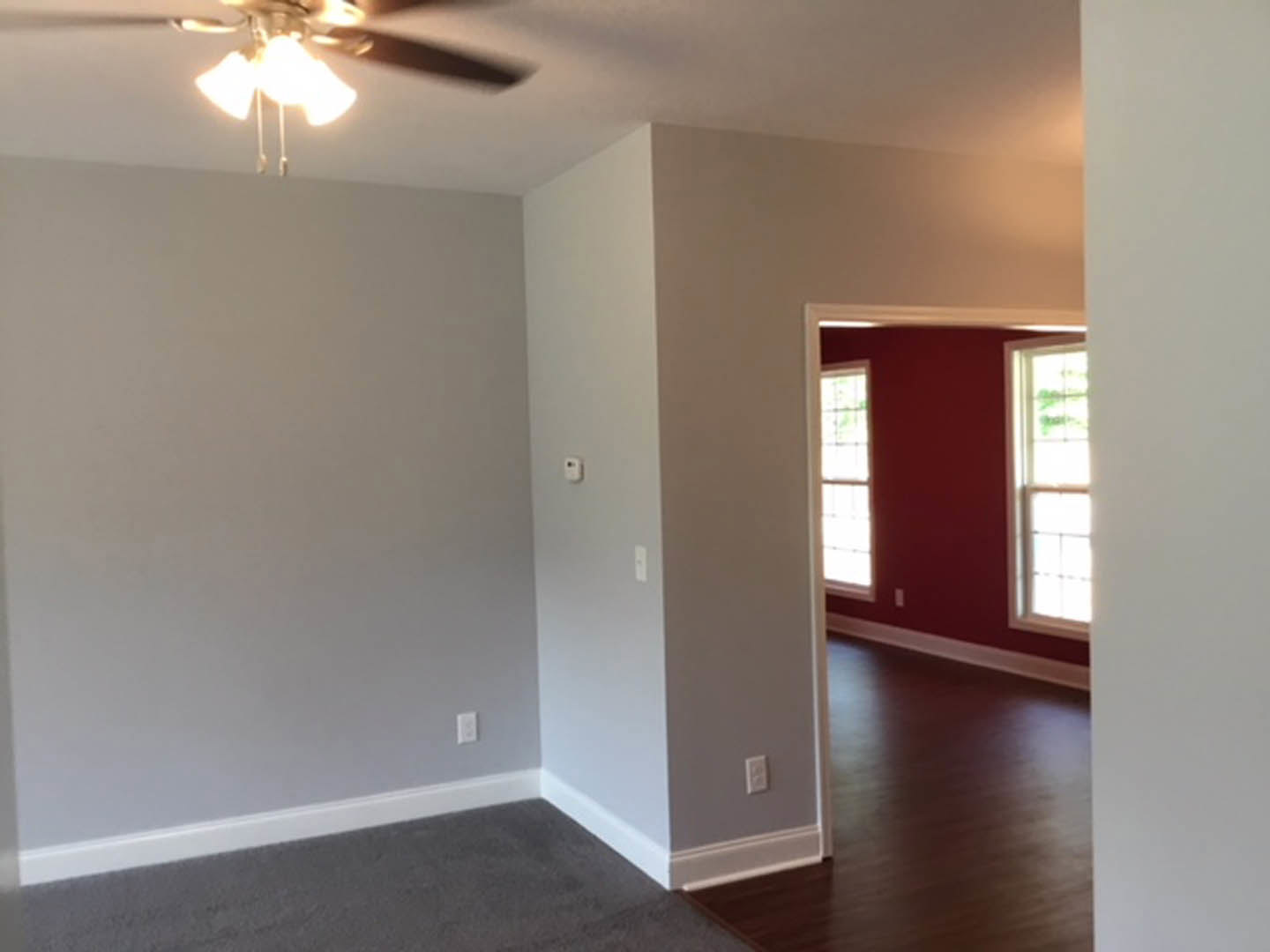 Room with dark wood flooring, white baseboard trim, red accent wall featuring windows, ceiling fan with light fixture, and green leafy plant visible through window.