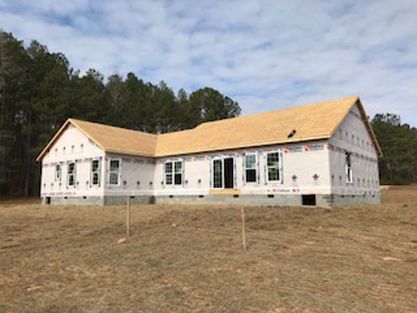 Partially built house with brown roof, exposed framing, fenced perimeter, patchy brown grass, surrounding trees, and overcast sky
