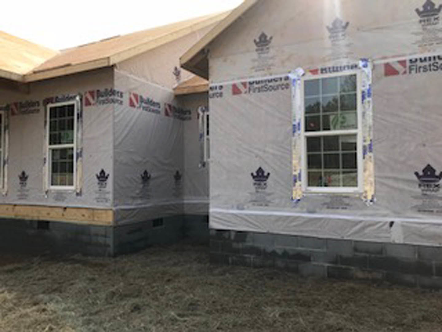 White custom home exterior with plastic sheeting draped over the facade, multi-pane window with white frame, black crown detail above, brick wall base, grassy lawn, and blurred