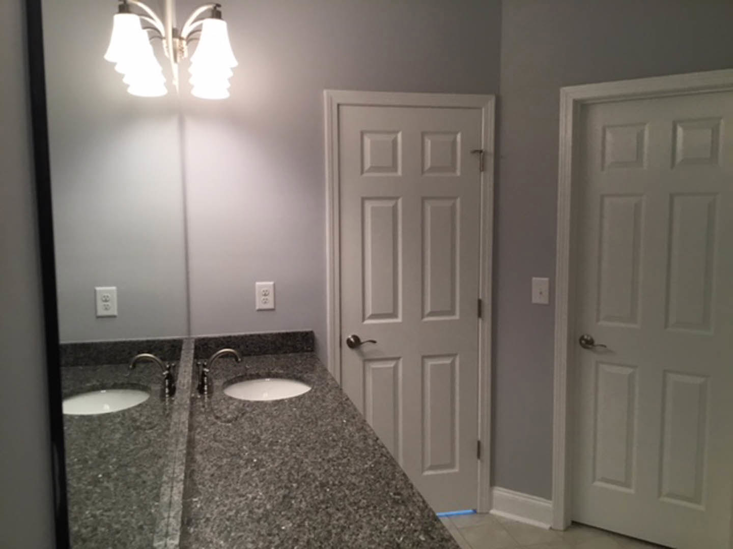 Bathroom with two undermount sinks set in a stone countertop, dual mirrors above, white cabinetry, tile flooring, and paneled doors with metal handles.