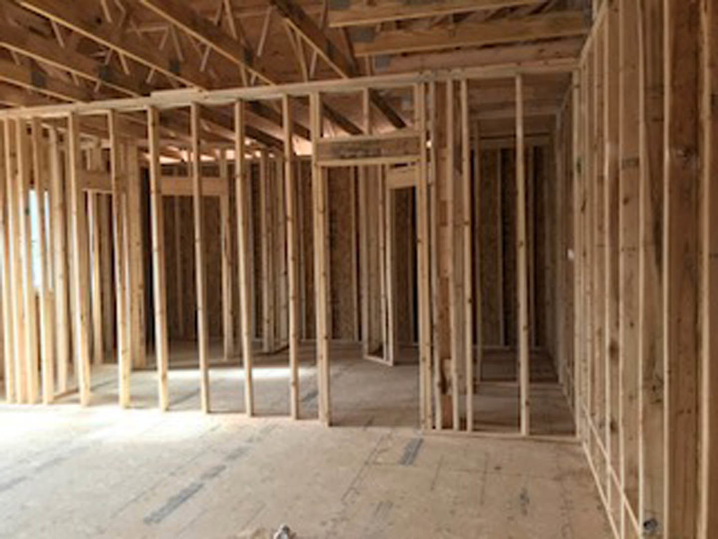Wood-framed room under construction with exposed ceiling beams, unfinished wooden floor, and partially visible window and hand in the background