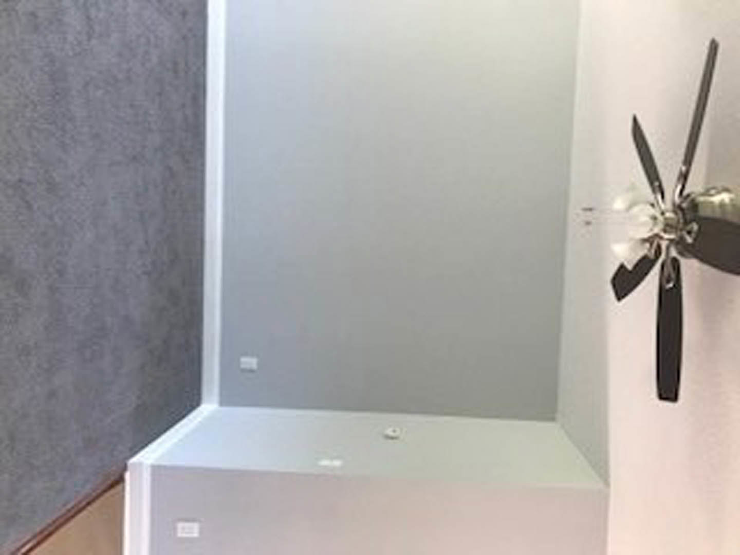 Ceiling fan with white blades mounted on a gray ceiling in a corner of a modern residential room, white square vent visible nearby.
