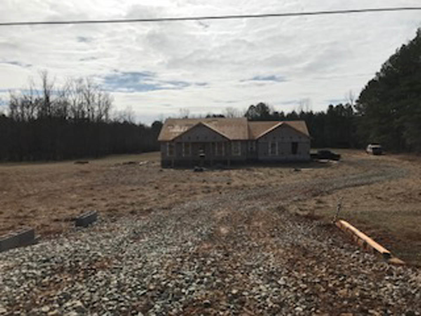 Gravel driveway leading to a cottage-style house surrounded by mature trees, wooden beam lying on the ground, power lines overhead, cloudy sky, grassy rural landscape