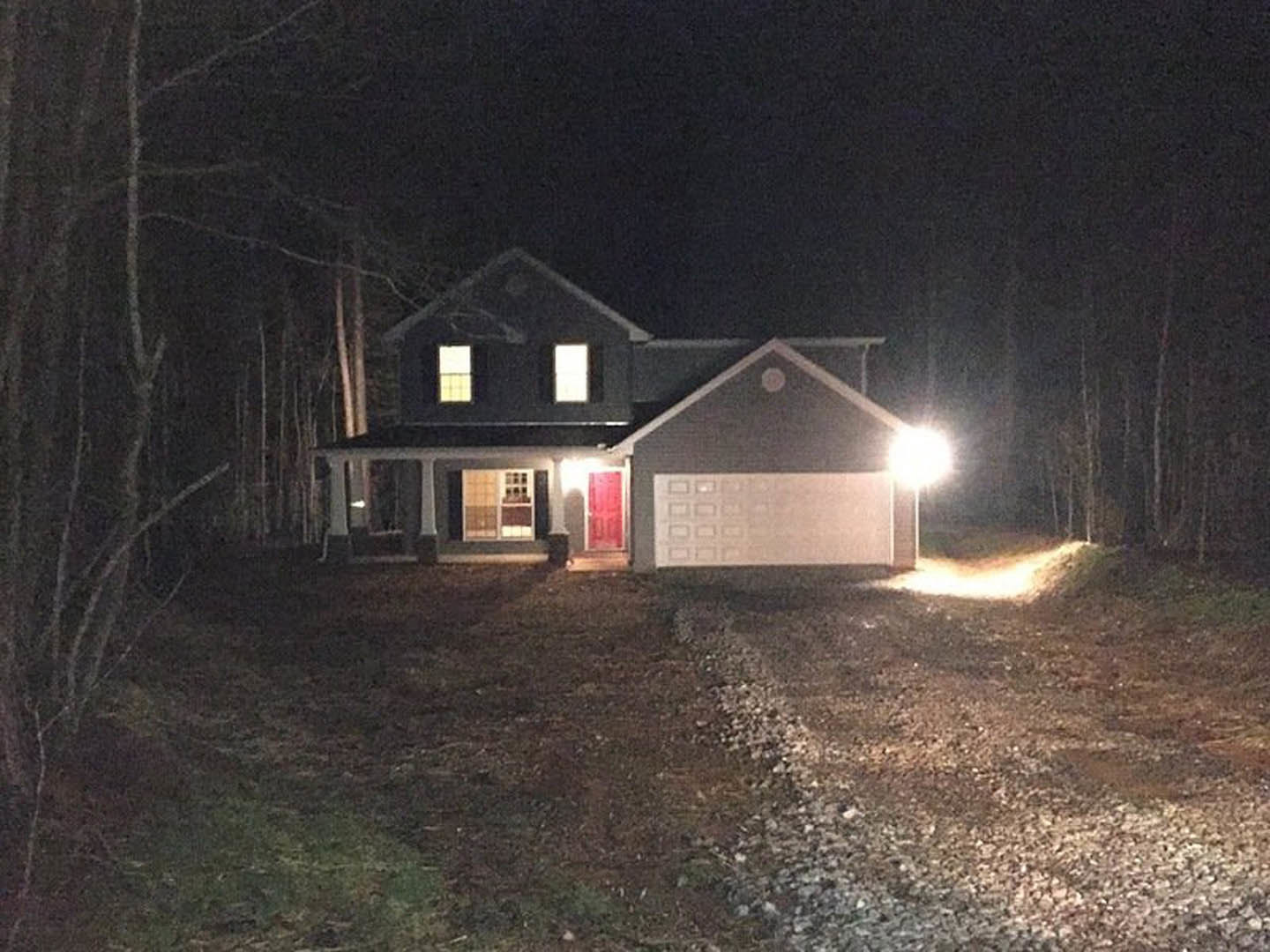 Two-story home with illuminated windows and exterior lights, red front door with white trim, attached garage, paved driveway, surrounding grass and trees at night