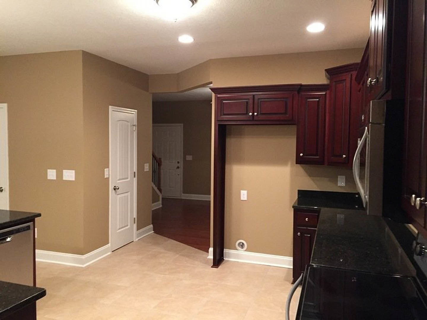 Dark wood kitchen cabinets with sleek black countertop, white door with silver knob, tile flooring, ceiling light fixture, wooden railing visible near entry.