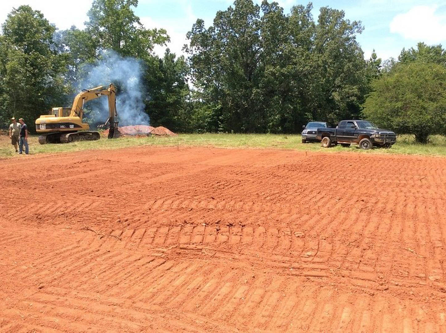 Black pickup truck and yellow excavator parked on a dirt field with tire tracks, surrounded by grass and trees under a cloudy sky