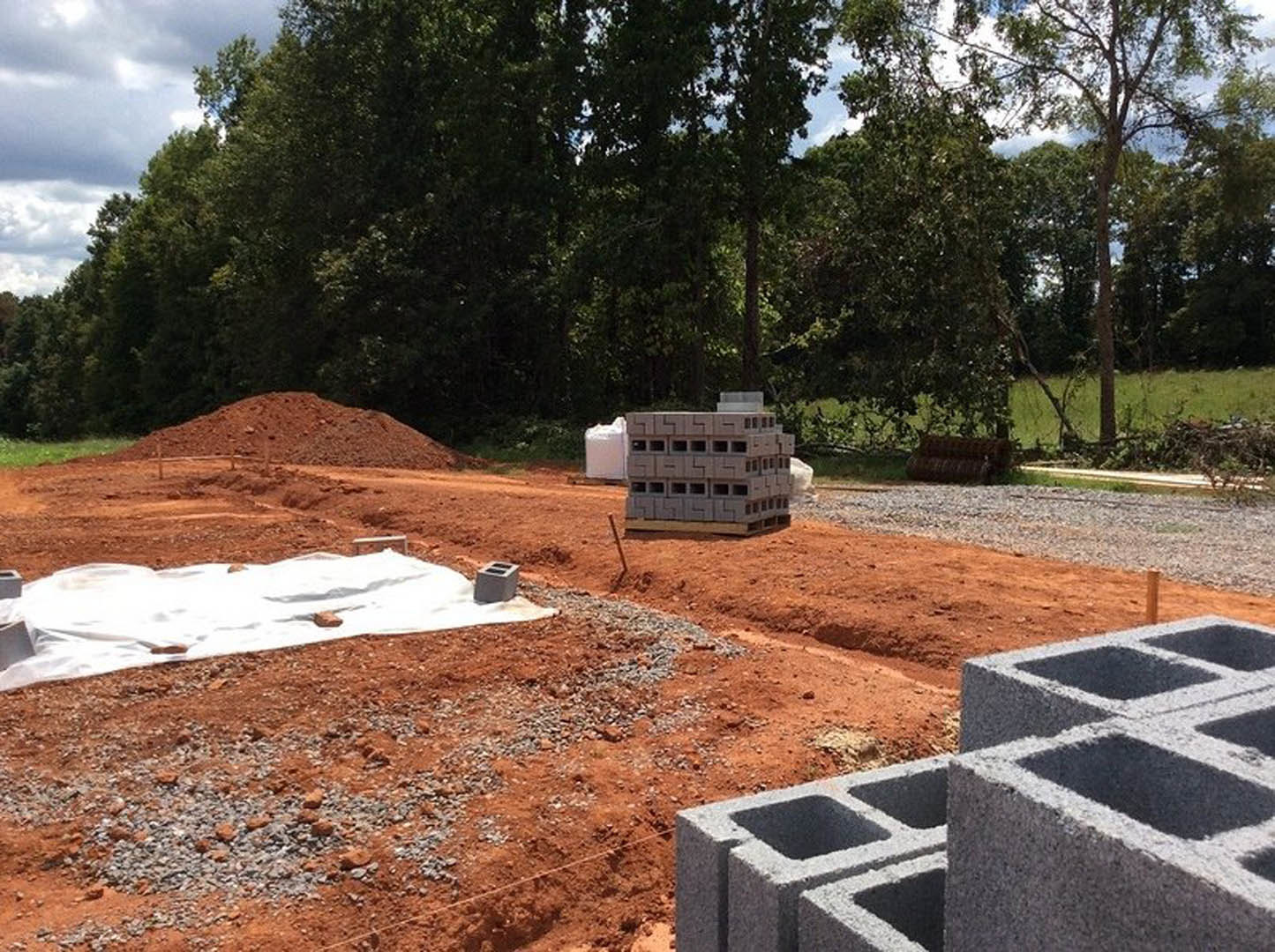 Construction site with stacked concrete blocks on dirt ground, surrounded by trees and cloudy sky