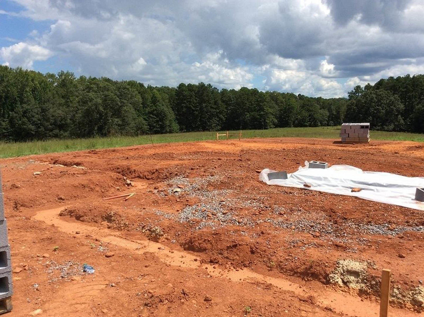 Dirt field with a white sheet spread on the ground, scattered bricks and a shovel nearby, cloudy sky and trees in the background