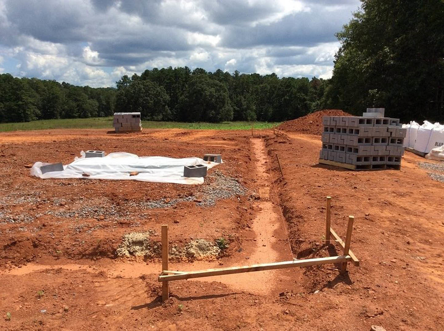 Cement blocks stacked on dirt at a residential construction site, wooden beam nearby, white cloth spread on ground, black object with square holes, cloudy sky and forest in