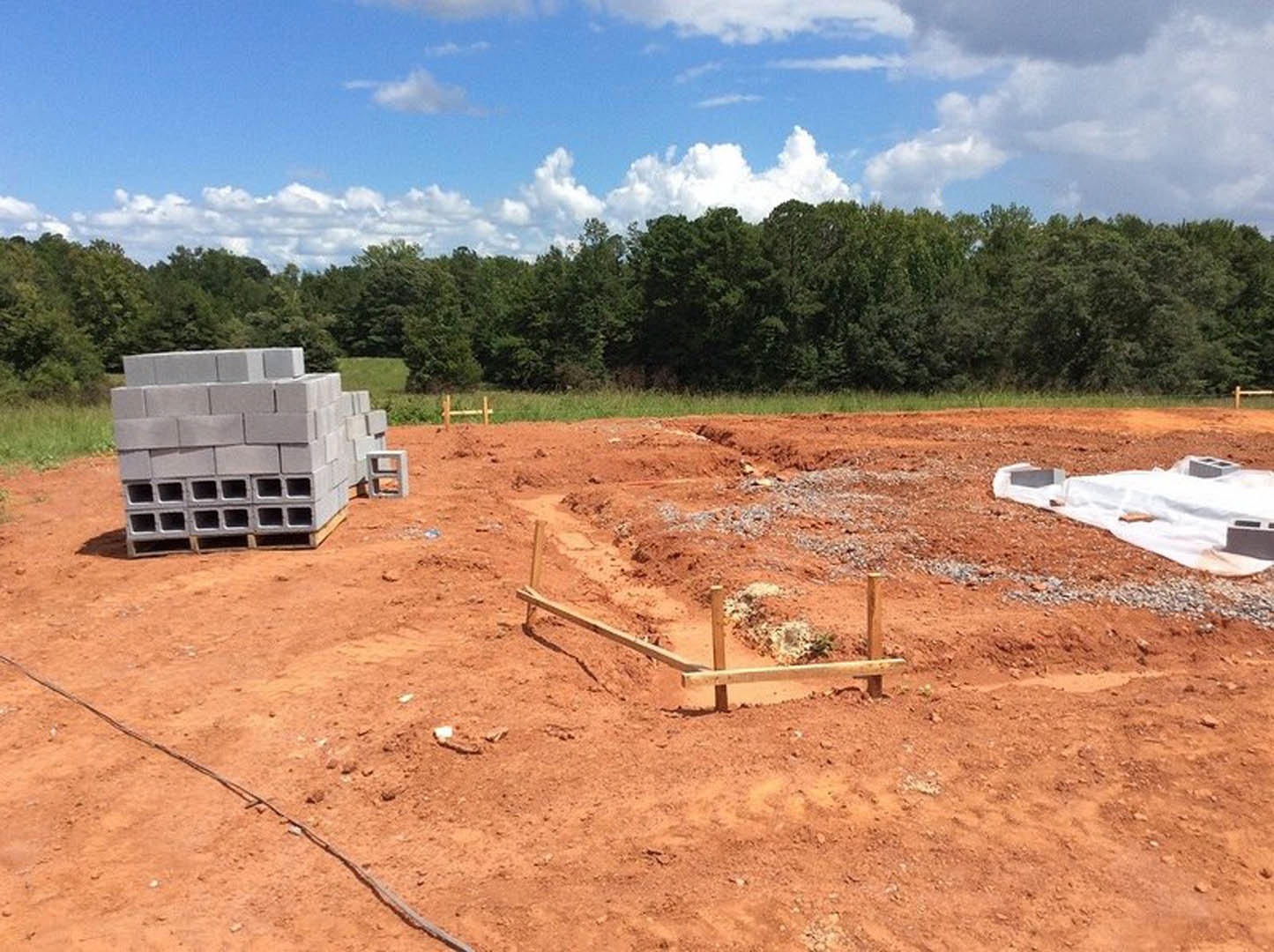 Stack of grey bricks beside a wooden fence on a dirt construction site, white sheet spread on the ground, trees in the background