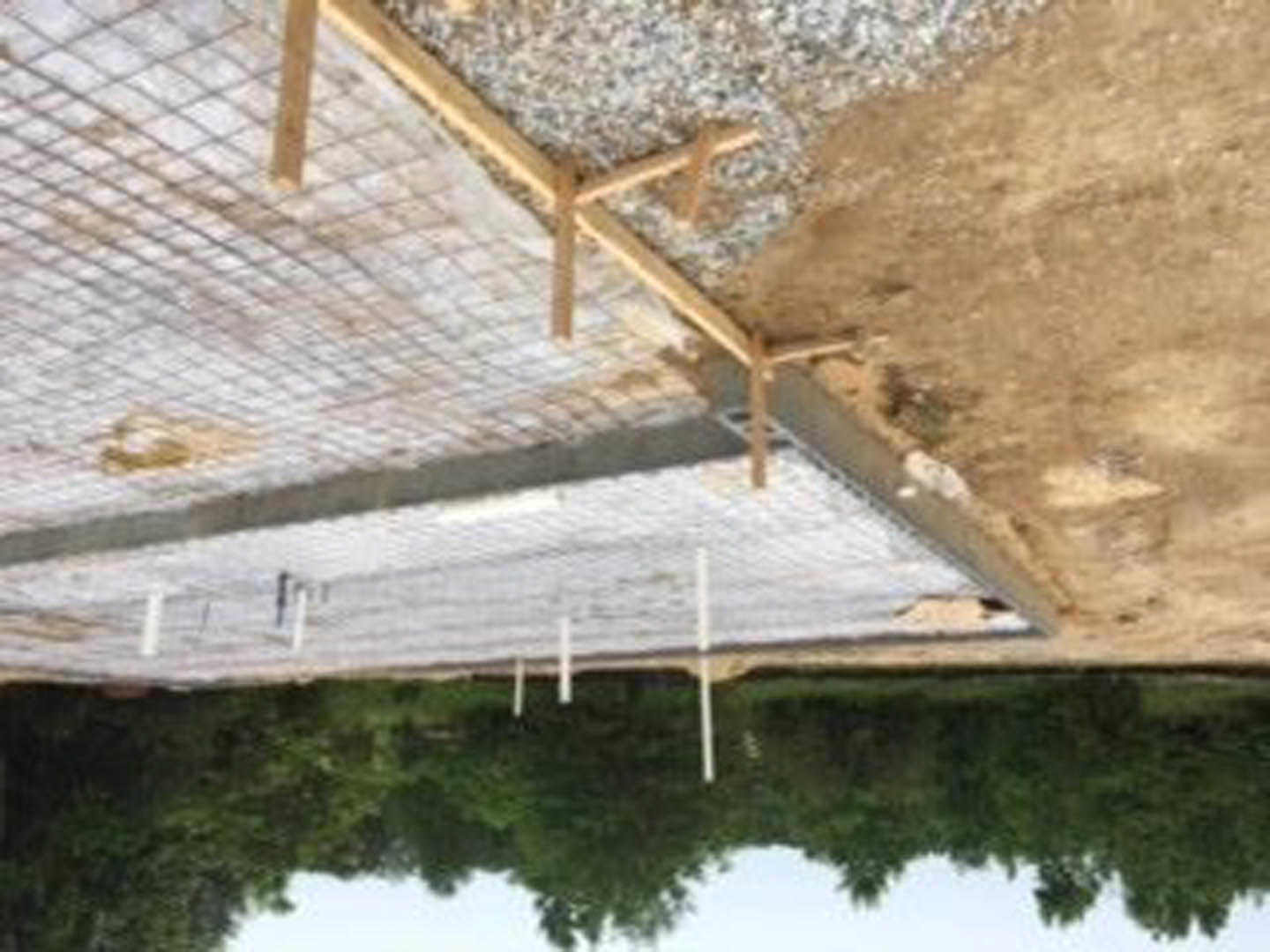 Wood framing and metal beams rising from a concrete foundation, surrounded by dirt and gravel, with mature trees in the background