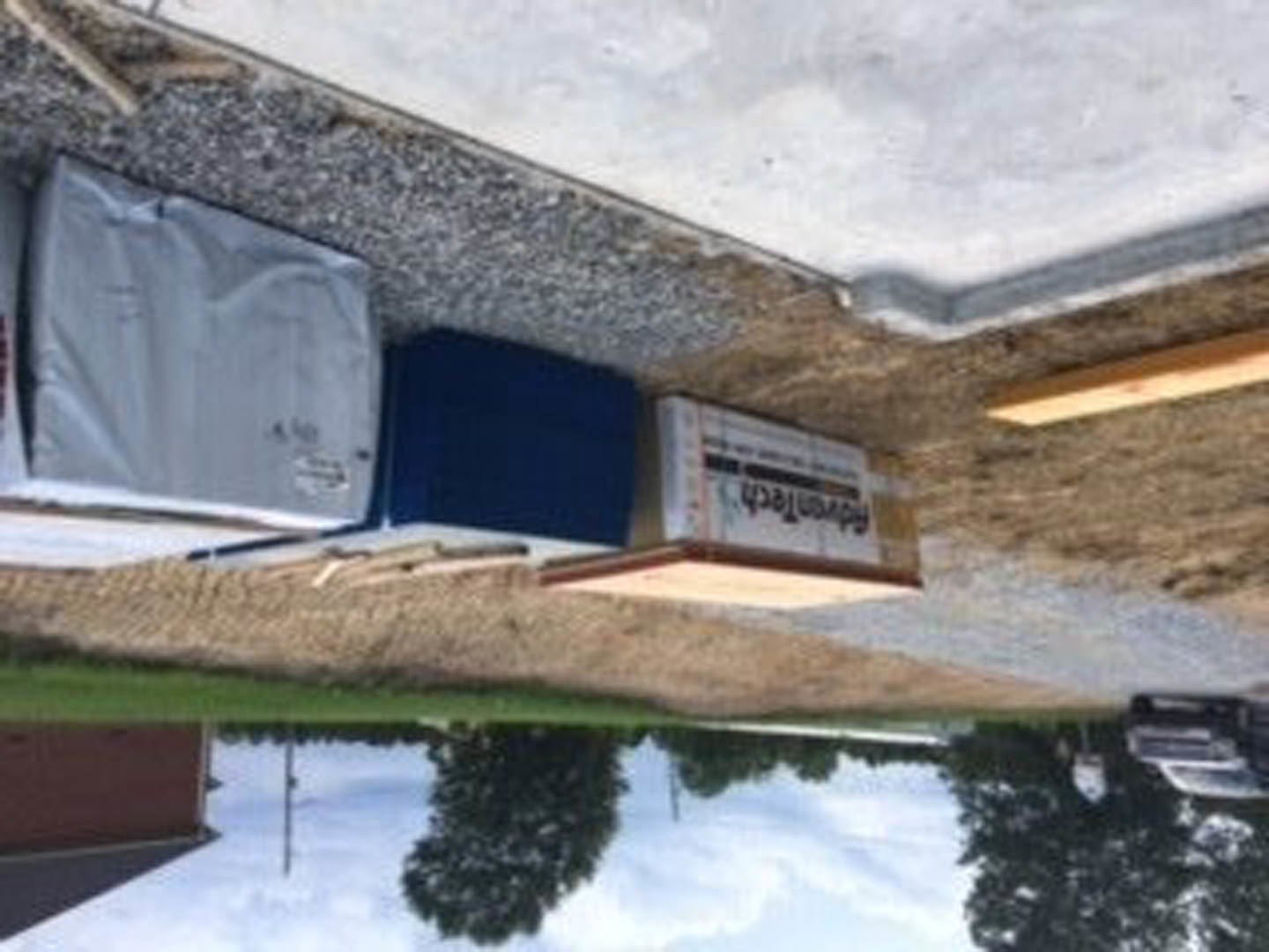 Cardboard boxes stacked beside a residential street, with a white square object on the pavement, a parked car, and a blue rectangular item near a house exterior; windows reflect