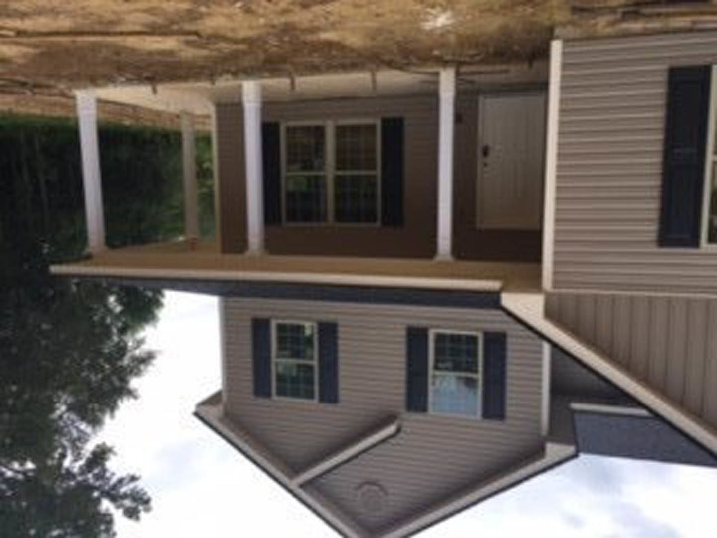 Large front porch with white railings, light-colored siding, multiple windows with white frames, and a prominent entry door; landscaped yard visible in foreground.