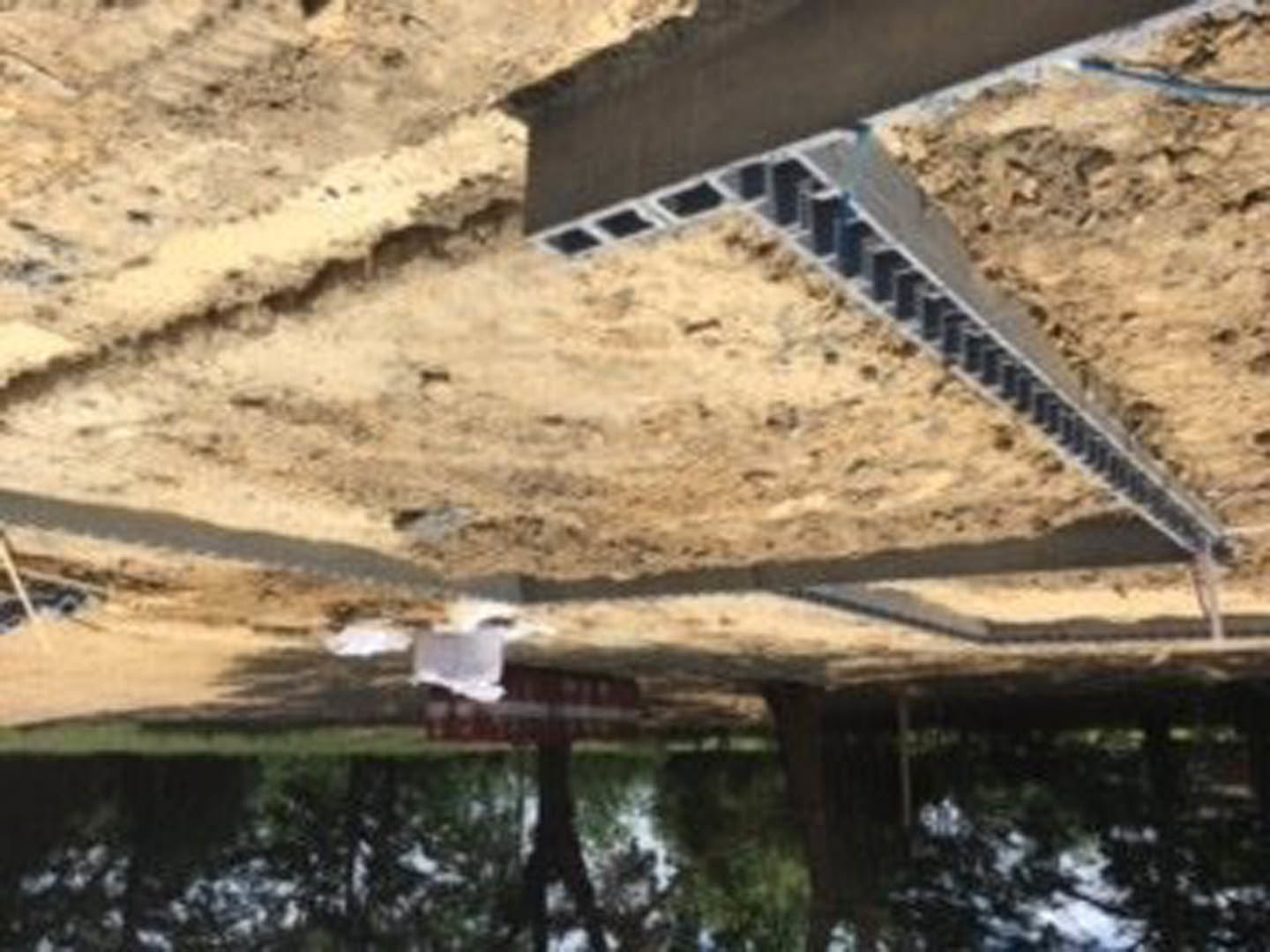 Concrete foundation with drilled holes beside a metal beam embedded in dirt, fenced boundary in background, tree visible, close-up of soil pile in foreground