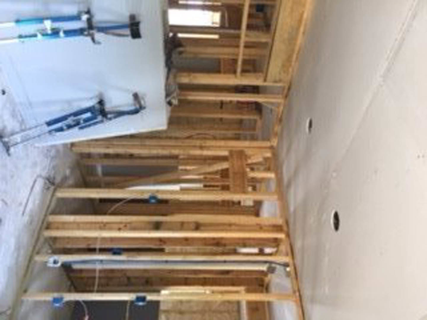 Exposed wood framing with plywood panels, blue and white crutches leaning against a white wall, unfinished interior construction with visible beams and insulation