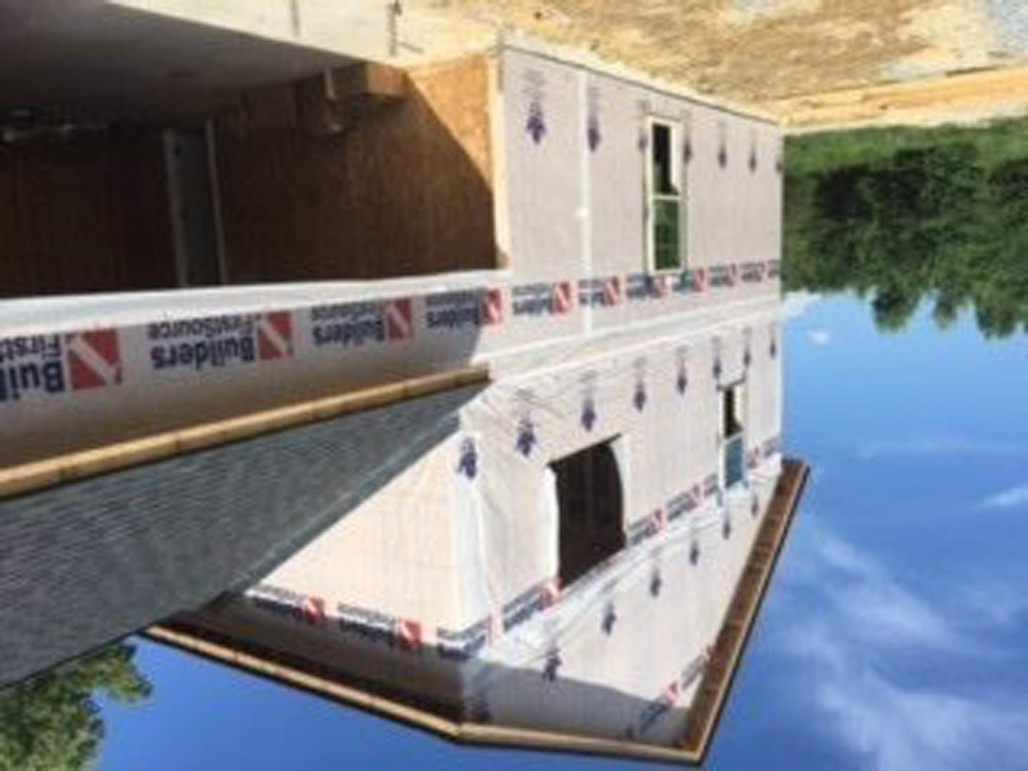 Modern home under construction with glass windows reflecting trees and blue sky, surrounded by greenery and water.