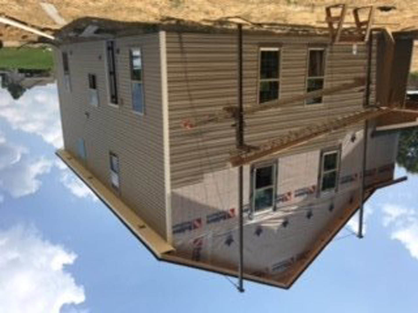 Partially built house with exposed framing, unfinished exterior walls, visible windows, and construction materials scattered around the site under a cloudy sky