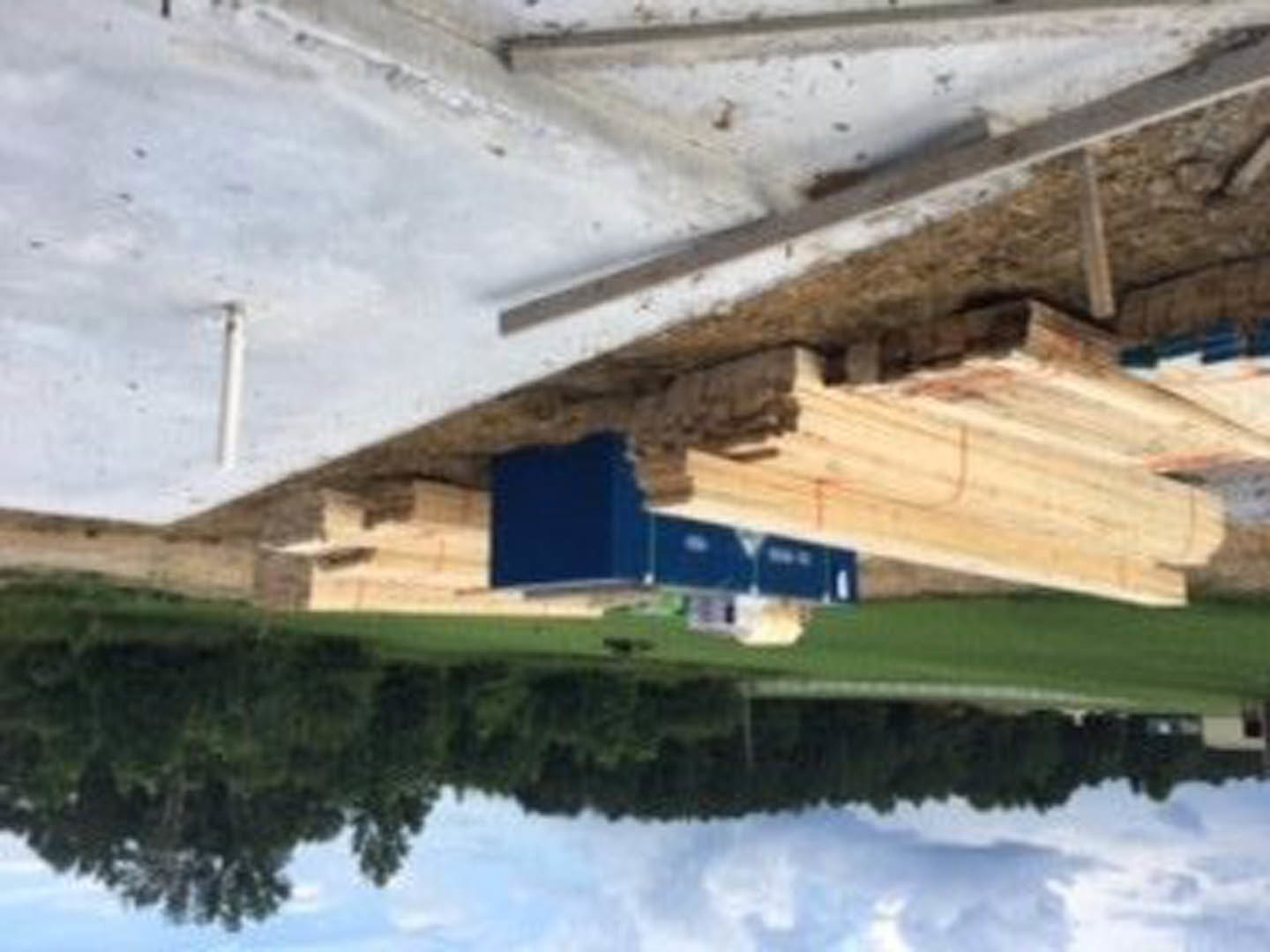 Stacked wooden boards beside a blue utility box on grassy ground, exterior of a modern house reflected in nearby water.