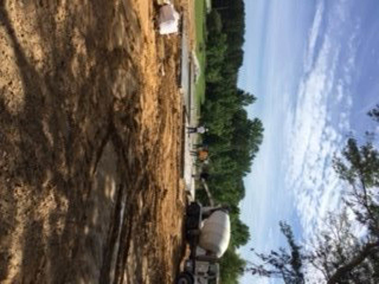 Cement mixer on a construction site with tree shadows on the ground, white container, blue sky with scattered clouds, and trees in the background