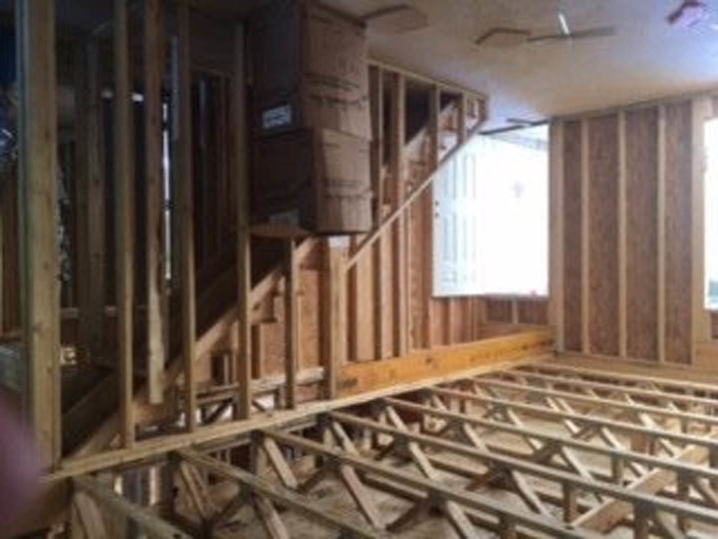 Open living space featuring two staircases, exposed wooden ceiling beams, light wood flooring, white door with decorative cross detail, and partial view of a wooden wall frame