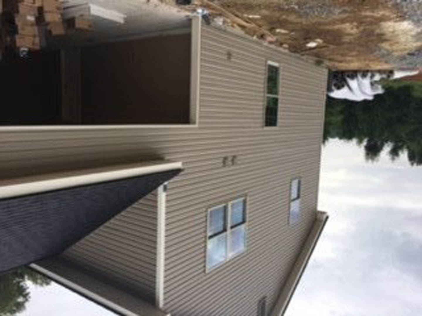 Partially built house with black roof, exposed framing, and construction materials, situated near a body of water, surrounded by trees and fencing