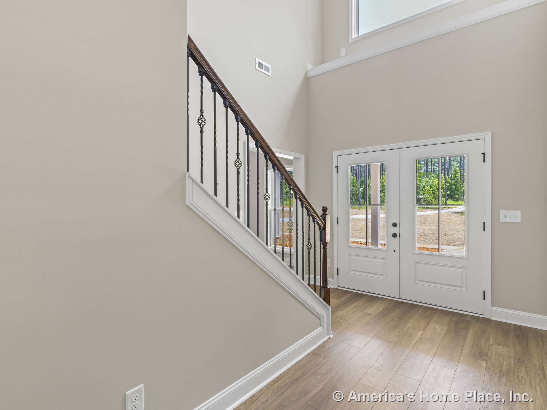 Double glass entry doors open to a foyer with light wood plank flooring, a wood staircase featuring iron balusters, large transom window above the doors, neutral wall paint, white