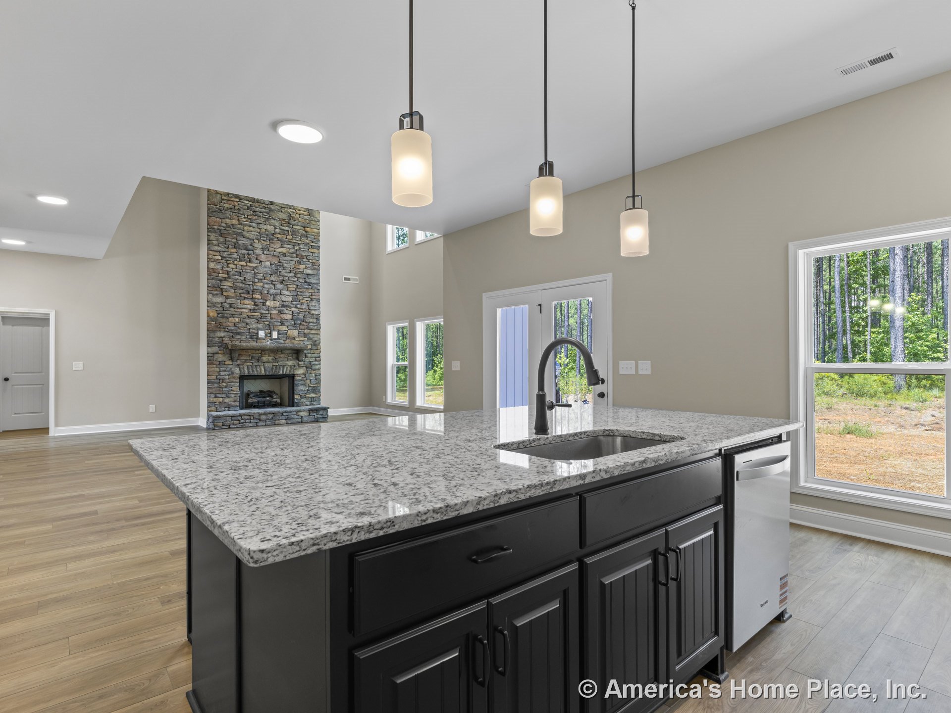 Open-concept living area with stone fireplace and mantel, expansive window wall with white trim, kitchen island featuring granite countertop, built-in sink, black cabinetry