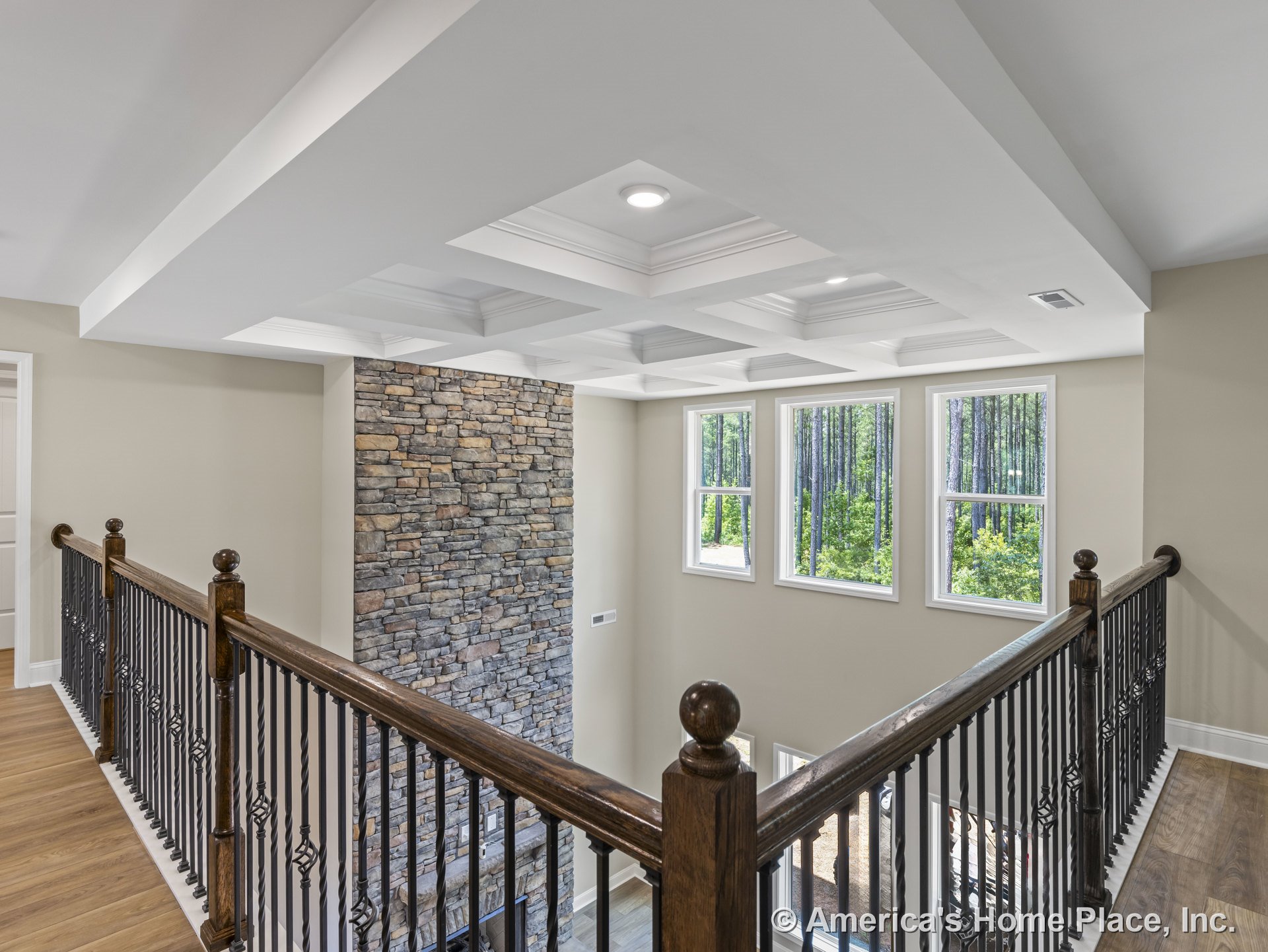 Upstairs landing with coffered ceiling and recessed lighting, stone accent wall, three large windows providing natural light and wooded views, wood and iron railing, light wood