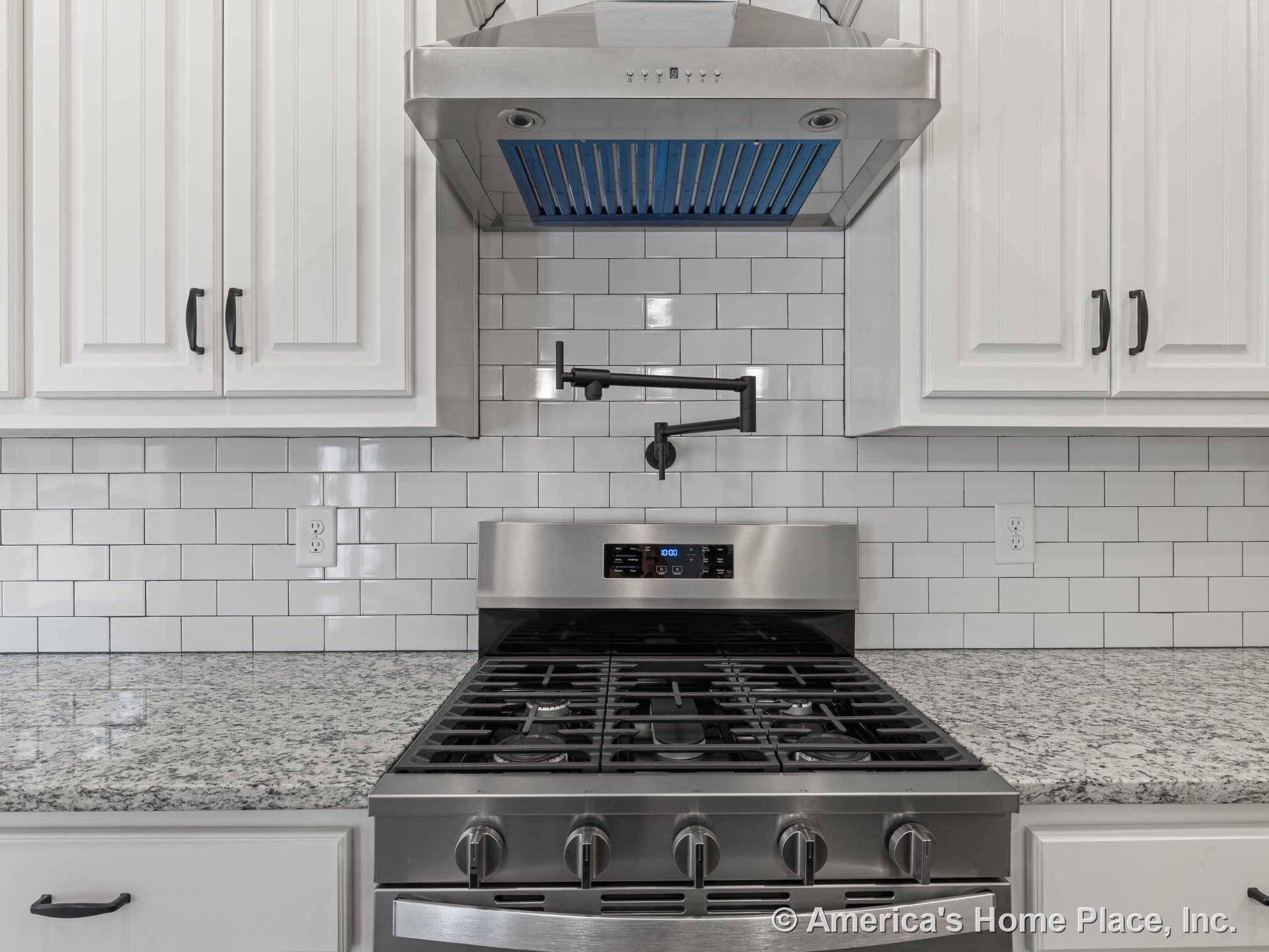 Stainless steel gas range with pot filler and vent hood featuring blue filter, granite countertops, white subway tile backsplash, white cabinetry with black hardware, built-in