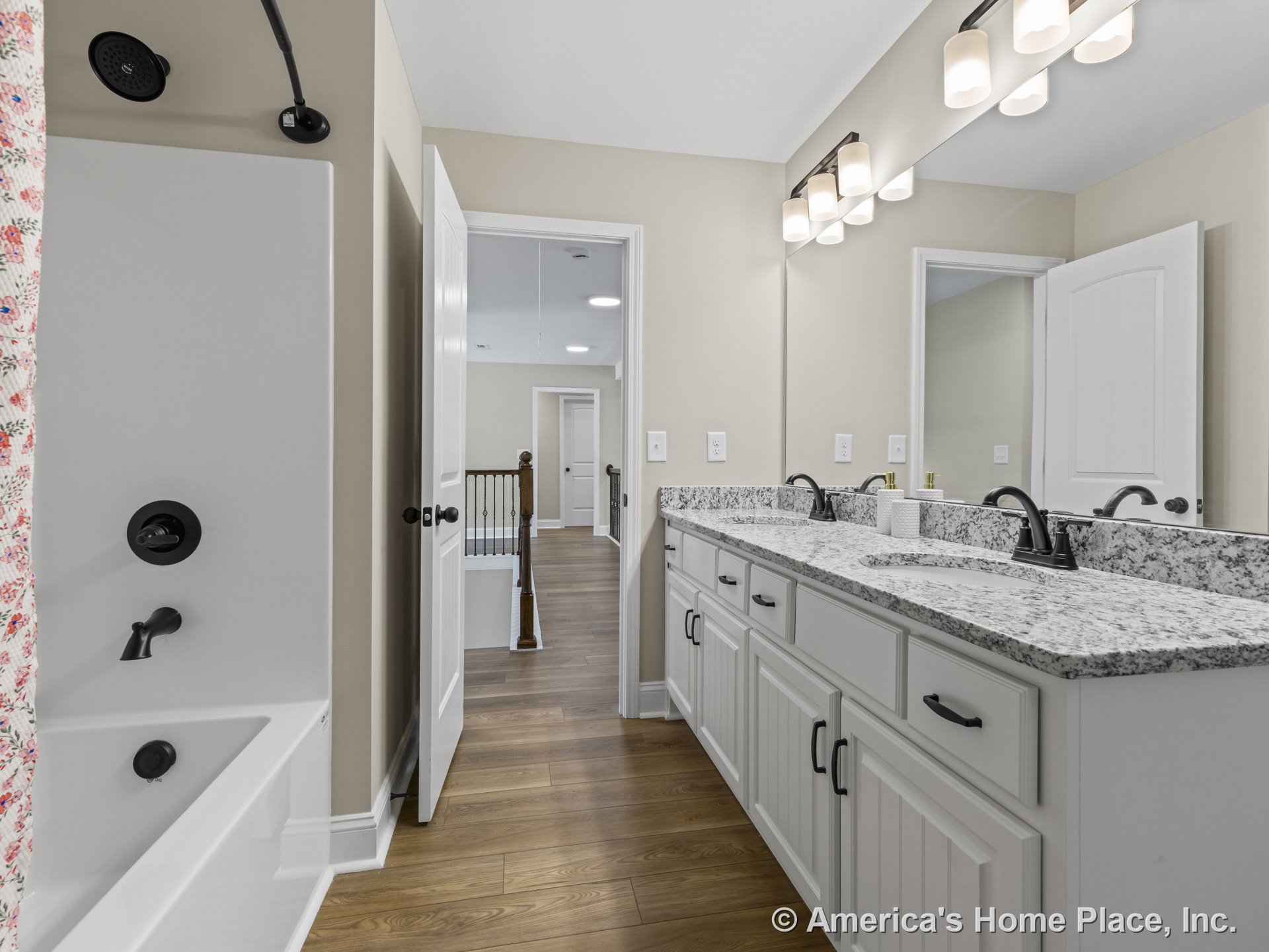 Double vanity with granite countertop, white shaker cabinets, black hardware, wood-look flooring, wall-mounted light fixtures above large mirrors, separate bathtub and