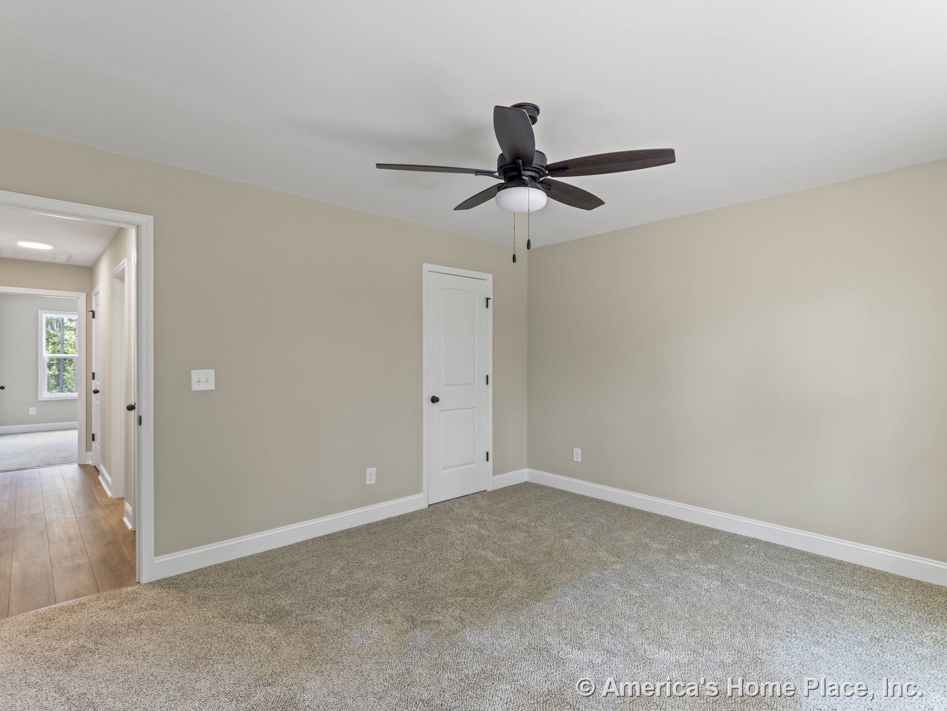 Bedroom with beige painted walls, carpet flooring, white trim and baseboards, ceiling fan with light fixture, white paneled door, and adjacent hallway featuring wood flooring.